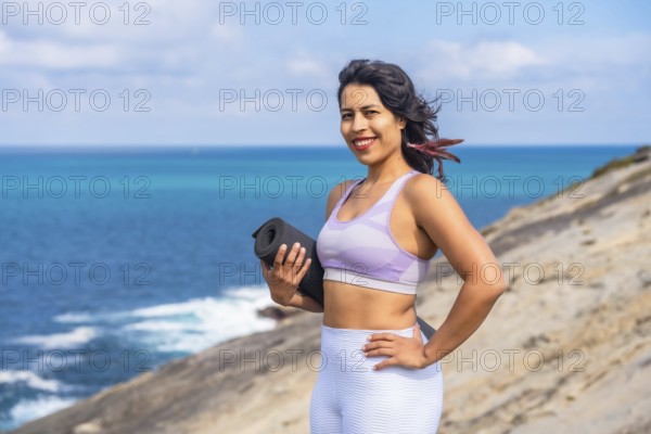 Woman smiling and holding a rolled yoga mat, standing confidently on a coastal cliff overlooking
