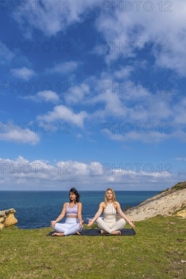 Two women practicing meditation and yoga in a serene lotus pose on a yoga mat overlooking the ocean