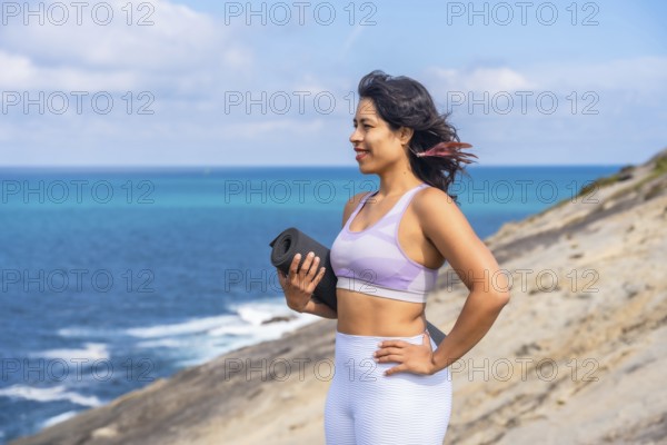 Woman standing on cliff by the ocean, holding a yoga mat and looking to the side, embodying