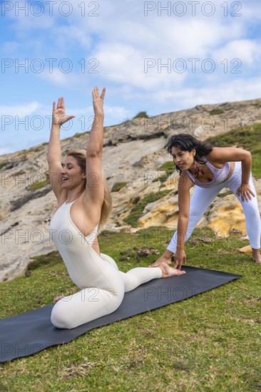 Two women practicing yoga together outdoors on a green grassy hill, one woman demonstrating a