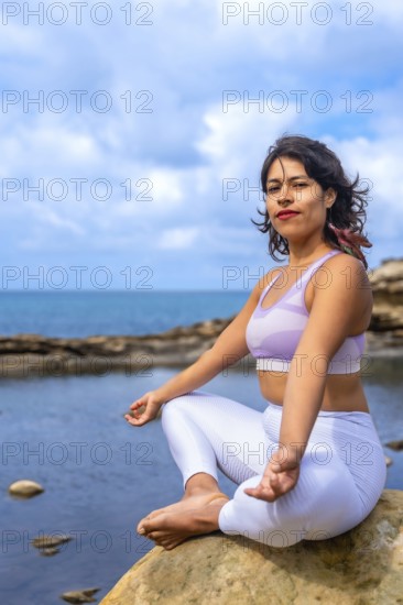 Woman sitting in a lotus position on a rock by the ocean, practicing yoga and finding inner peace,