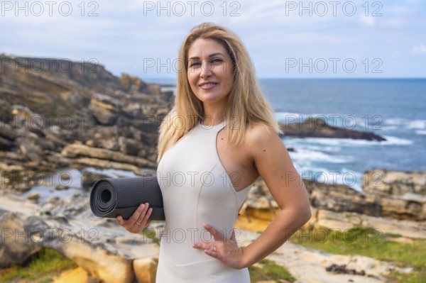 Woman stands on rocky coast by the ocean holding a yoga mat, smiling at camera before a yoga and