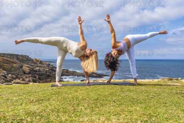 Two women performing a balancing yoga pose on mats on a grassy cliff overlooking the ocean,