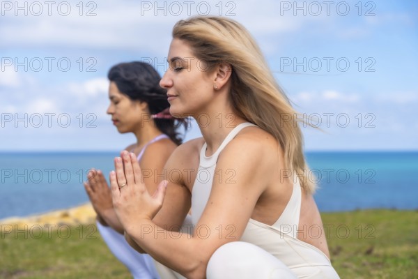 Two women with eyes closed practicing yoga and meditation in prayer pose on the seaside, embracing