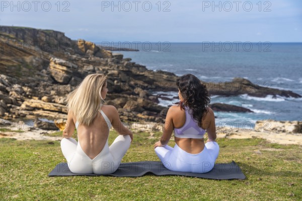 Two women practicing yoga and meditation together on a mat, sitting outdoors on a green cliff