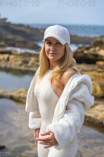 Woman in white sportswear and baseball cap stands on rocky shoreline by the blue sea, smiling