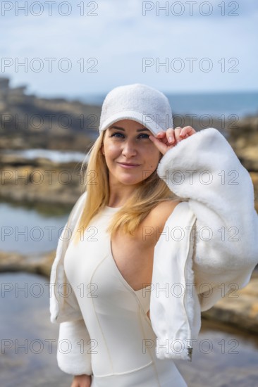Young woman wearing a white cap and dress, standing confidently on rocky coastal terrain, with the
