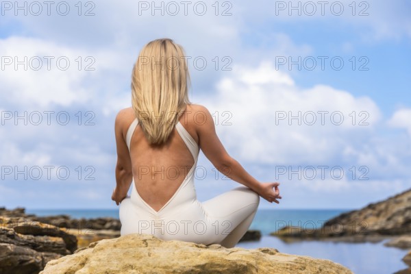 Woman in a white jumpsuit sitting in a peaceful meditation pose on an ocean cliff, finding balance