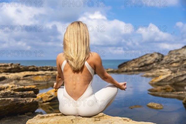 Woman sitting facing the ocean practicing yoga in a meditative pose on coastal rocks, finding