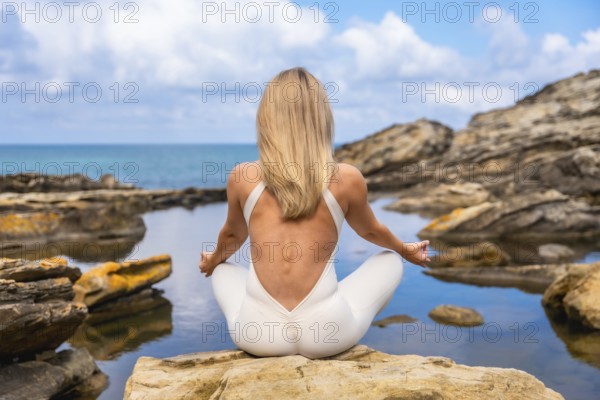 Woman in a white jumpsuit practicing a meditation yoga pose on coastal rocks, finding wellness and