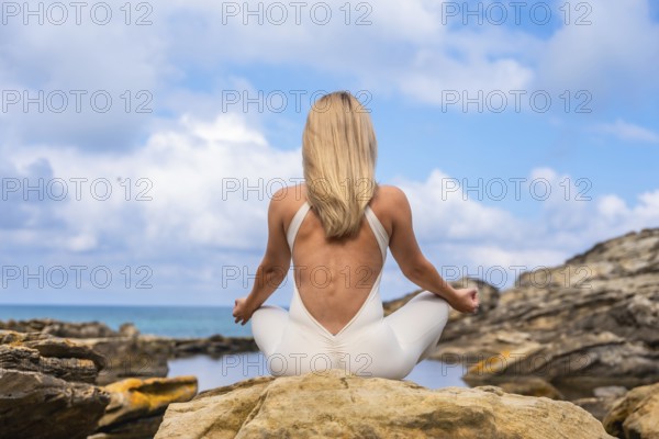 Woman in yoga attire seated in lotus on a coastal rock, meditating with calm focus amid blue ocean