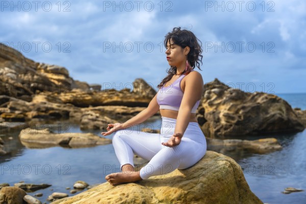 Woman sitting in a yoga pose, meditating outdoors on a rock by the sea, finding tranquility and