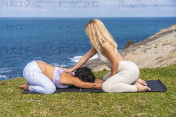 Two women are practicing yoga on a mat on a grassy cliff overlooking the ocean, with one woman