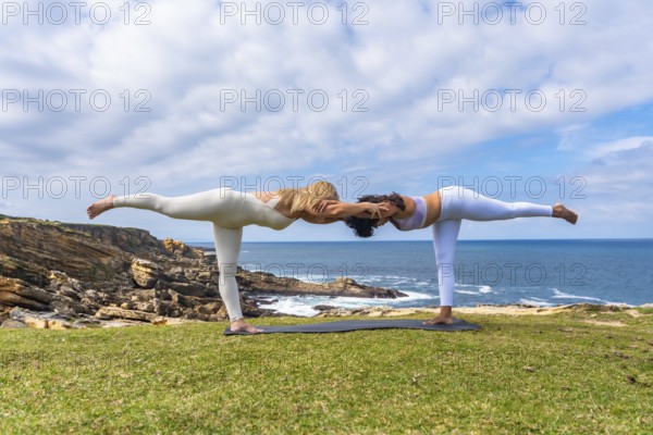 Two women practicing acro yoga outdoors on a grassy cliff overlooking the ocean, holding a deep