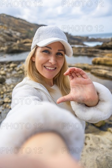 Blonde woman in white cap and jacket smiling at camera on a rocky beach, forming a heart with her