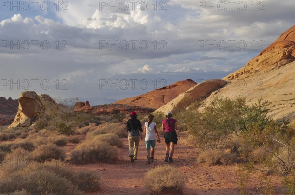 Three hikers on a trail through a desert landscape with clouds, Valley of Fire State Park, Nevada,