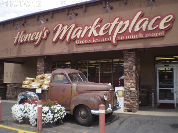Old Studebaker pickup truck full of flowers parked in front of a nostalgic market building, Honey's