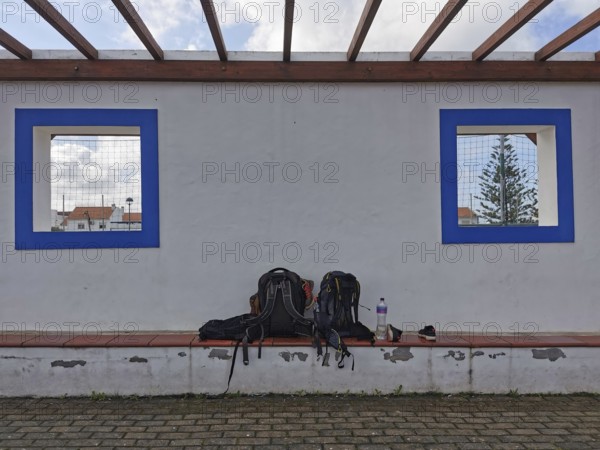 Two backpacks on a bench in front of a white wall with blue window cutouts, hiking on the