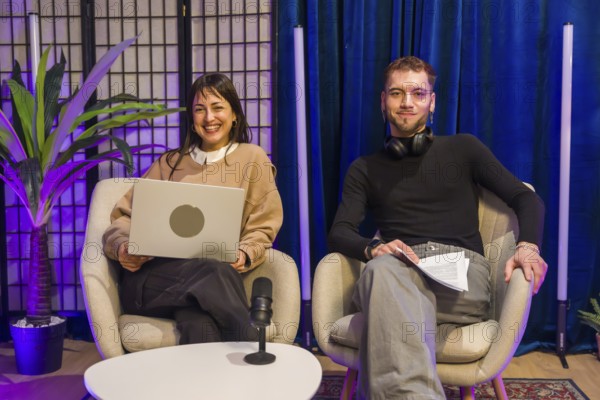Two smiling content creators, a woman holding a laptop and a man holding notes, sitting in chairs