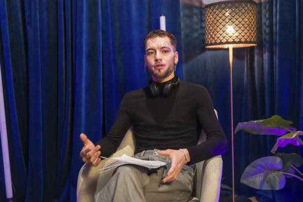 Young man wearing headphones around his neck, holding papers, and gesturing while speaking,