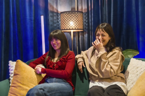 Two young women sitting on a sofa, one smiling and the other laughing, collaborating creatively in