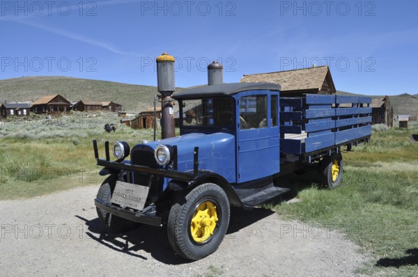 A blue historic truck with yellow wheels, Dodge Graham 1927, stands in front of a wooden house