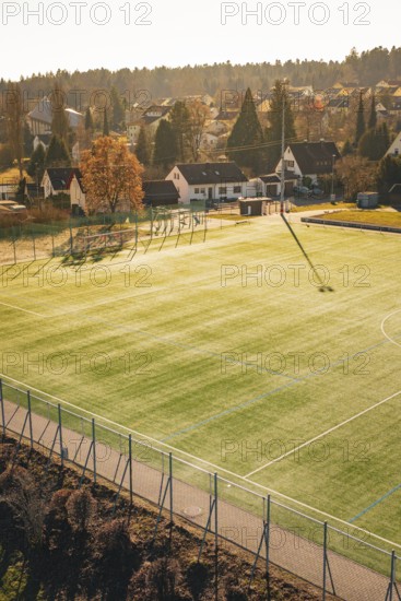 Sunlight illuminates a soccer field in the village with houses and trees in the background,