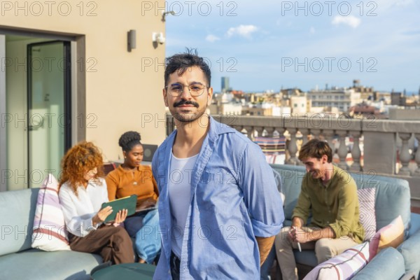 Smiling man standing on a rooftop terrace as diverse colleagues relax and work in the background,