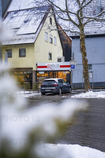 Snowy city street with parked car in front of a building with a shop, Gechingen, Calw district,