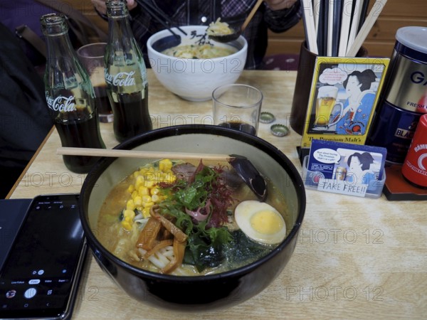 A bowl of ramen with egg, corn and vegetables on a table in a restaurant, accompanied by cola
