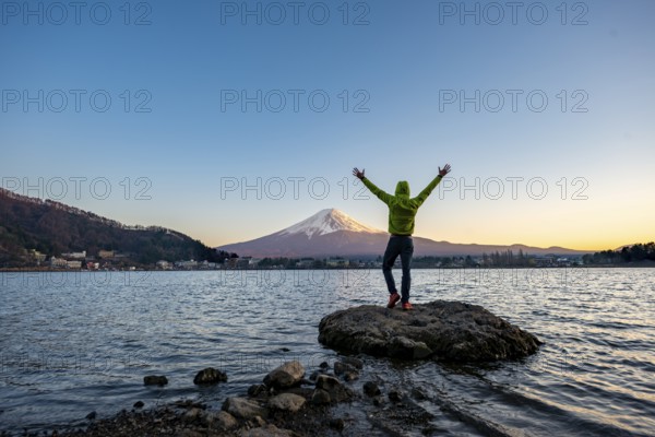 Young man stretching his arms in the air, standing on rocks in Lake Kawaguchi, view of Mount Fuji