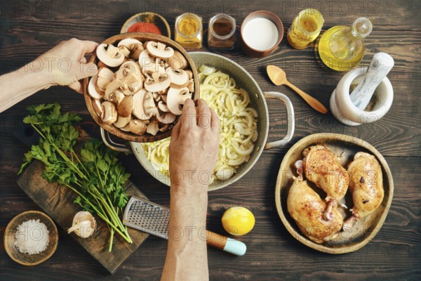 Two hands hold a bowl of sliced mushrooms over a pot with cooking onions on a wooden table.