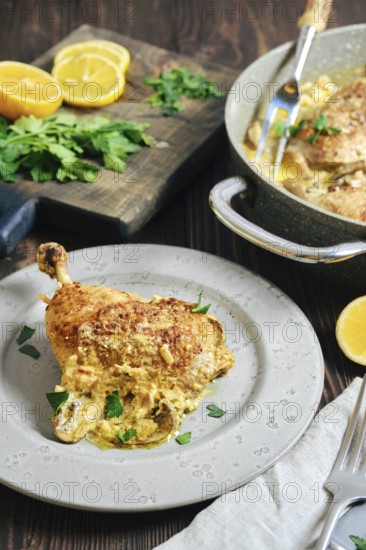 A chicken portion on a plate, surrounded by lemon slices and herbs. Cooking pan with more chicken