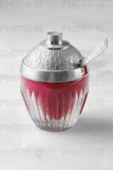 A bowl made of glass with a silver lid is filled with red lingonberry jam or sauce and sits on a