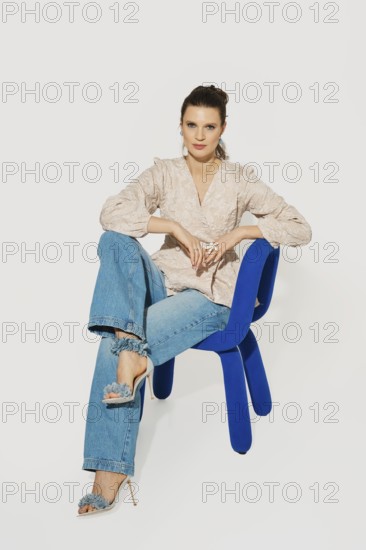 A woman sits on a blue chair with one leg crossed over the other. She wears a light top and denim