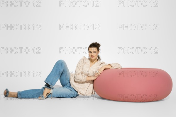 A woman in a light top and blue jeans is sitting next to a large pink cushion. She rests her arm on
