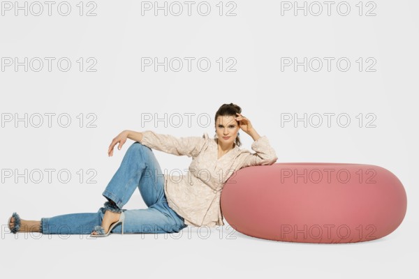 Cute woman is sitting on the floor beside a round pink chair in a studio. She is wearing a light
