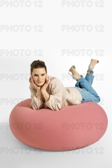 A woman lays on a big pink cushion in a studio. She is wearing a light blazer and jeans. Her hair