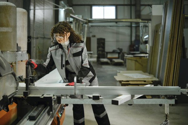 Woman is focused on using a large circular saw in a factory. The worker wears safety gear and