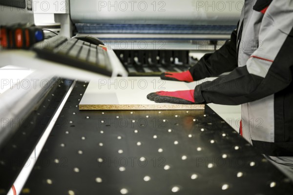 A person in gloves operates a machine that cuts a piece of material in a workshop. The workspace