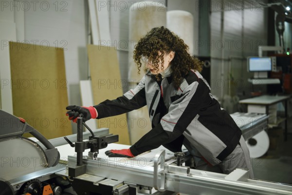 A woman in a workshop operates a table saw machine. She uses her hands to adjust a piece of wood.