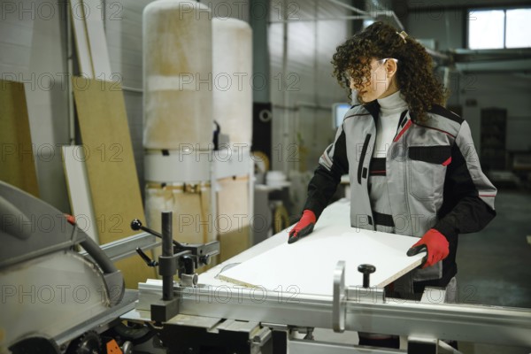 Female worker stands in a workshop focused on cutting a wood board with a table saw. The person
