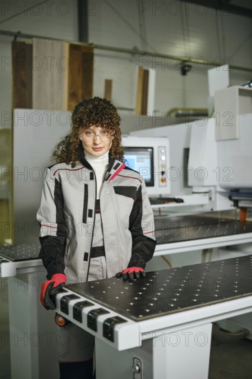 A woman stands in a workshop next to a wood shaping machine. She wears a work suit and gloves while