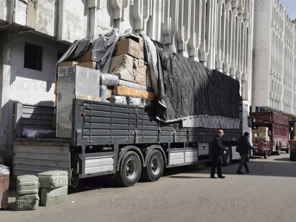 Large truck on the road, heavily loaded with boxes and covers, next to building, load securing,