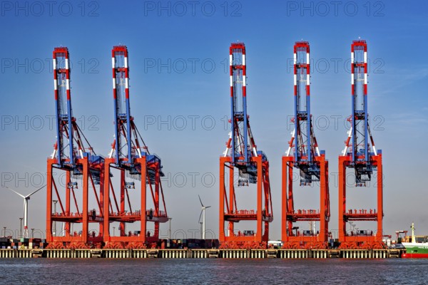 Set of red and blue cranes at the harbor, wind turbines and container warehouse in the background,