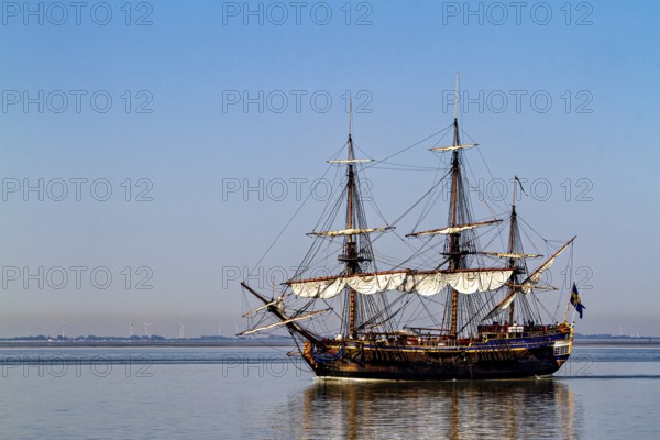 A traditional sailing ship with full sails on calm water under clear blue skies, an old historic