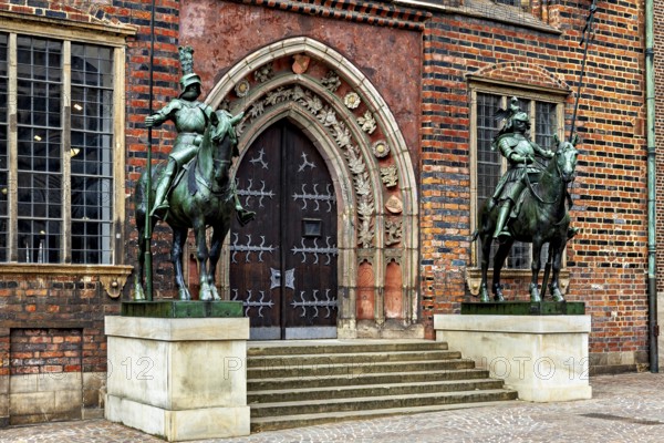 Two equestrian statues in front of a Gothic building with decorated entrance doors and red bricks,