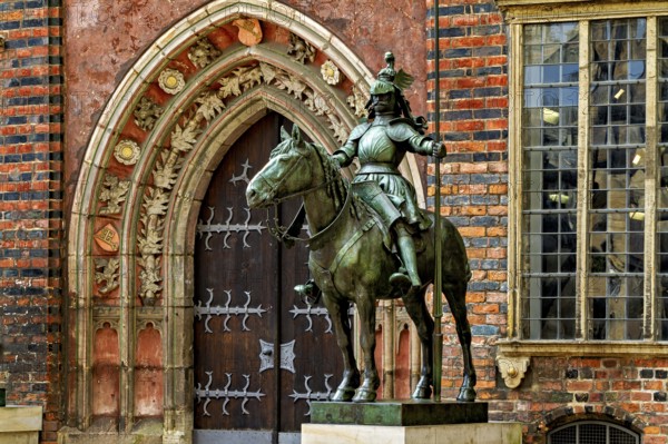 An equestrian statue in front of a Gothic building with richly decorated archway and red bricks,