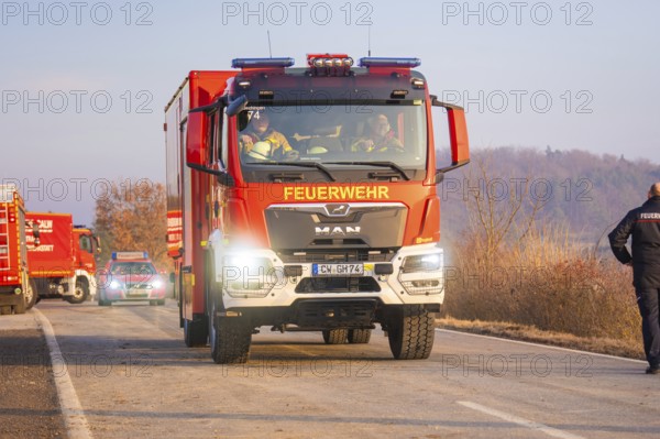 A fire truck drives on a road through a wooded area with emergency personnel, fire brigade exercise