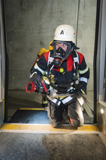 A firefighter wearing respiratory protection kneels in a tunnel at the threshold of a door, fire